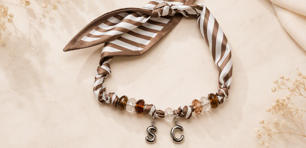 A beaded charm necklace with a brown and white striped fabric ribbon, brown beads, and silver letter charms spelling 'SC'.